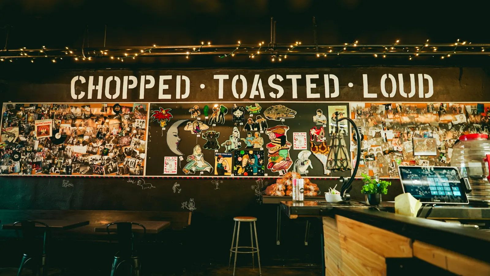 Inside the original NoshPit sandwich bar: a black wall stenciled with the words Chopped, Toasted, Loud above collages of band photos and handmade art